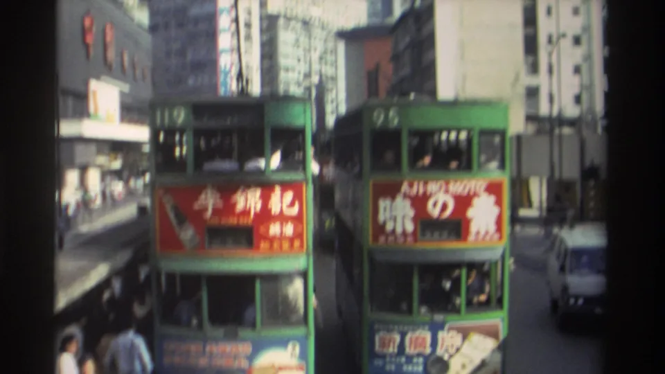 Green double-decker trams 119 and 95 on a Hong Kong street in 1982, Lee Kum Kee soy sauce and Aji-no-Moto advertisement panels visible, dense high-rise buildings behind, captured on 8mm film.