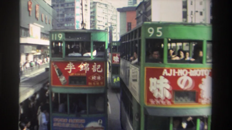 Hong Kong double-decker trams 119 and 95 in 1982, Lee Kum Kee and Aji-no-Moto livery panels prominent, upper-deck passengers seen through tram windows, dense urban high-rises in background.