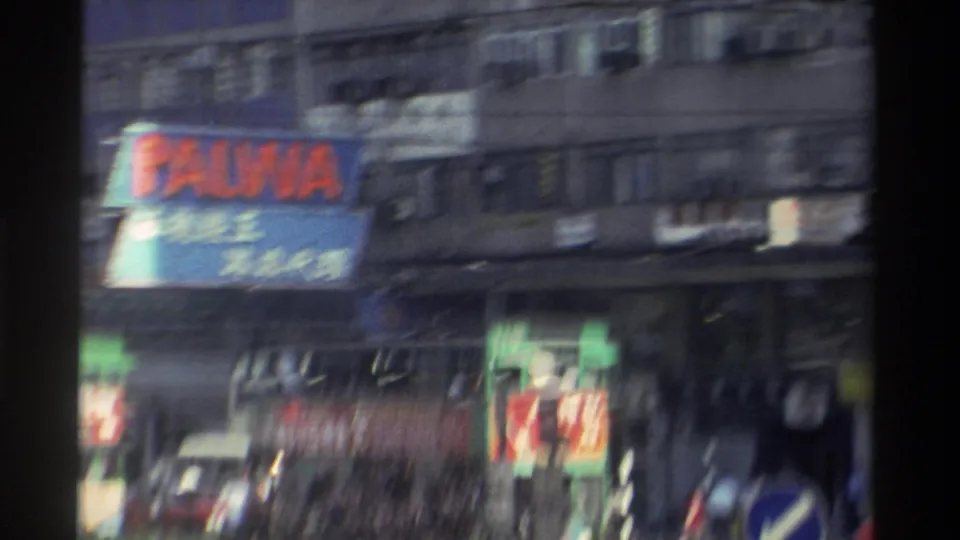 Close-angle 8mm frame from Hong Kong, 1982, with the PALWA billboard and stacked Chinese commercial signs; motion blur and grain are pronounced.