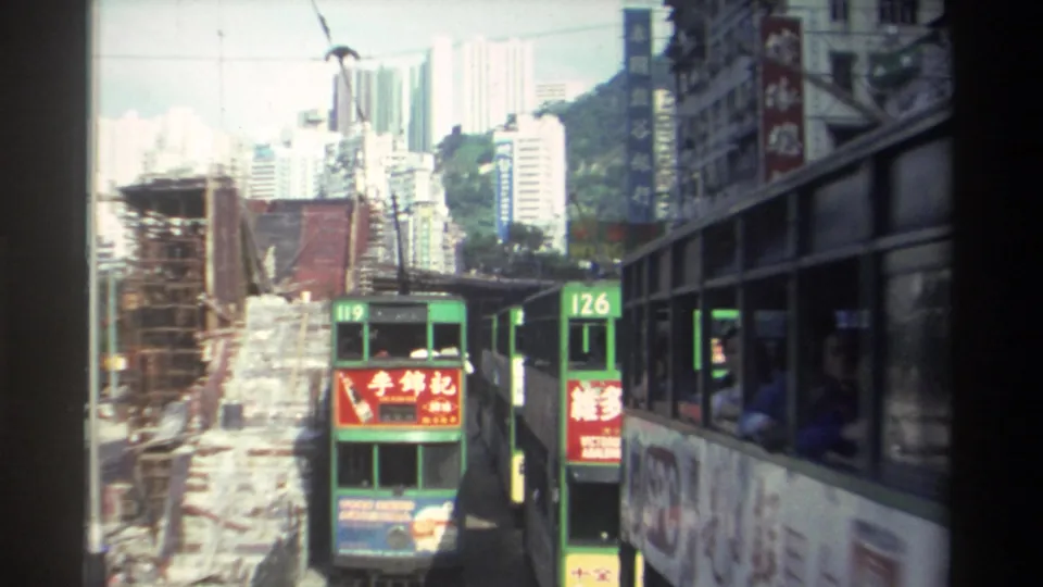 Steadier 8mm frame from Hong Kong, 1982, showing green double-decker trams numbered 119 and 126, bamboo-style construction scaffolding, residential high-rises, and overhead tram wires under a partly cloudy sky.