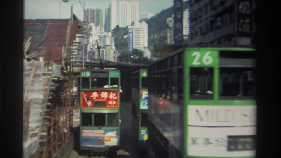 1982 Hong Kong 8mm frame showing tram 119 aligned with construction scaffolding as a green tram with a large white advertising panel passes close in the foreground.