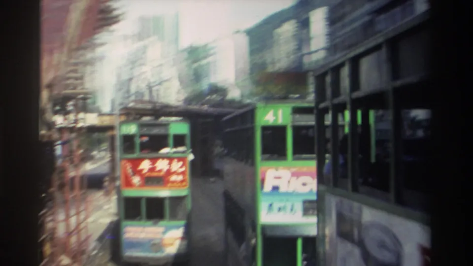 1982 Hong Kong home-movie frame with motion blur as green double-decker trams pass through a constricted street lined by scaffolding, advertising panels, buildings, and overhead wires.