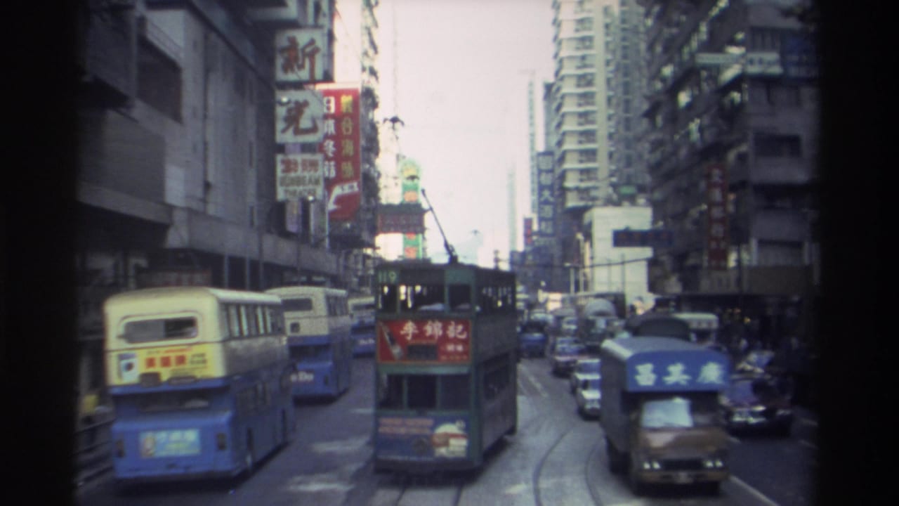1982 Hong Kong: blue-and-yellow double-decker bus and tram share a congested road lined with multi-storey buildings covered in Chinese commercial signage.