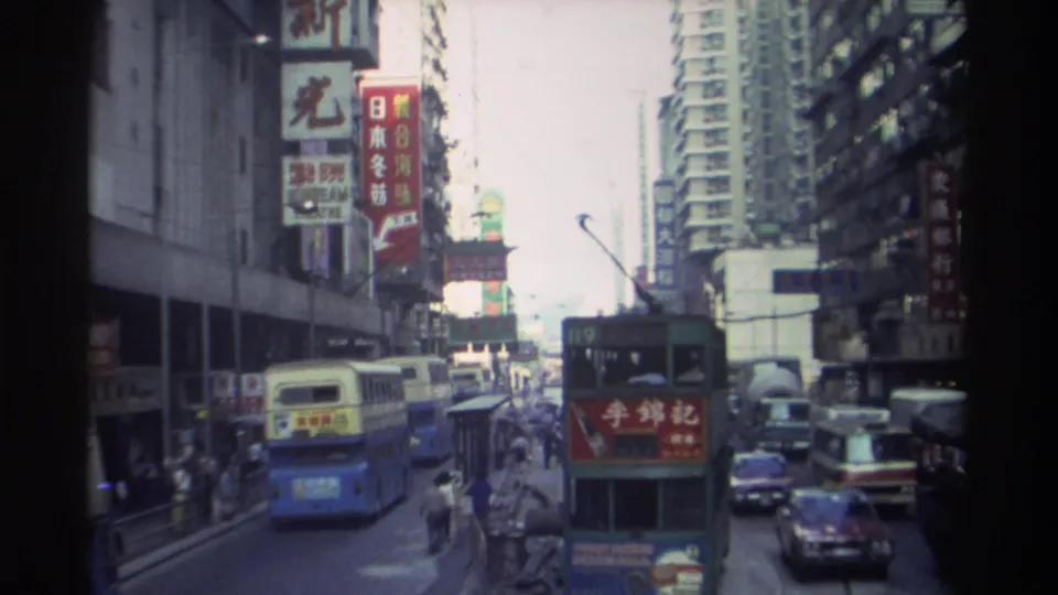 1982 Hong Kong: a double-decker bus with billboard advertising passes beneath layered commercial signage — including partial English-language text — on buildings lining a busy urban corridor.
