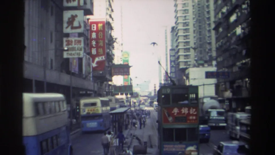 1982 Hong Kong: street-level view of double-decker buses and dense mixed-language signage amid tall commercial and residential buildings lining an active urban corridor.
