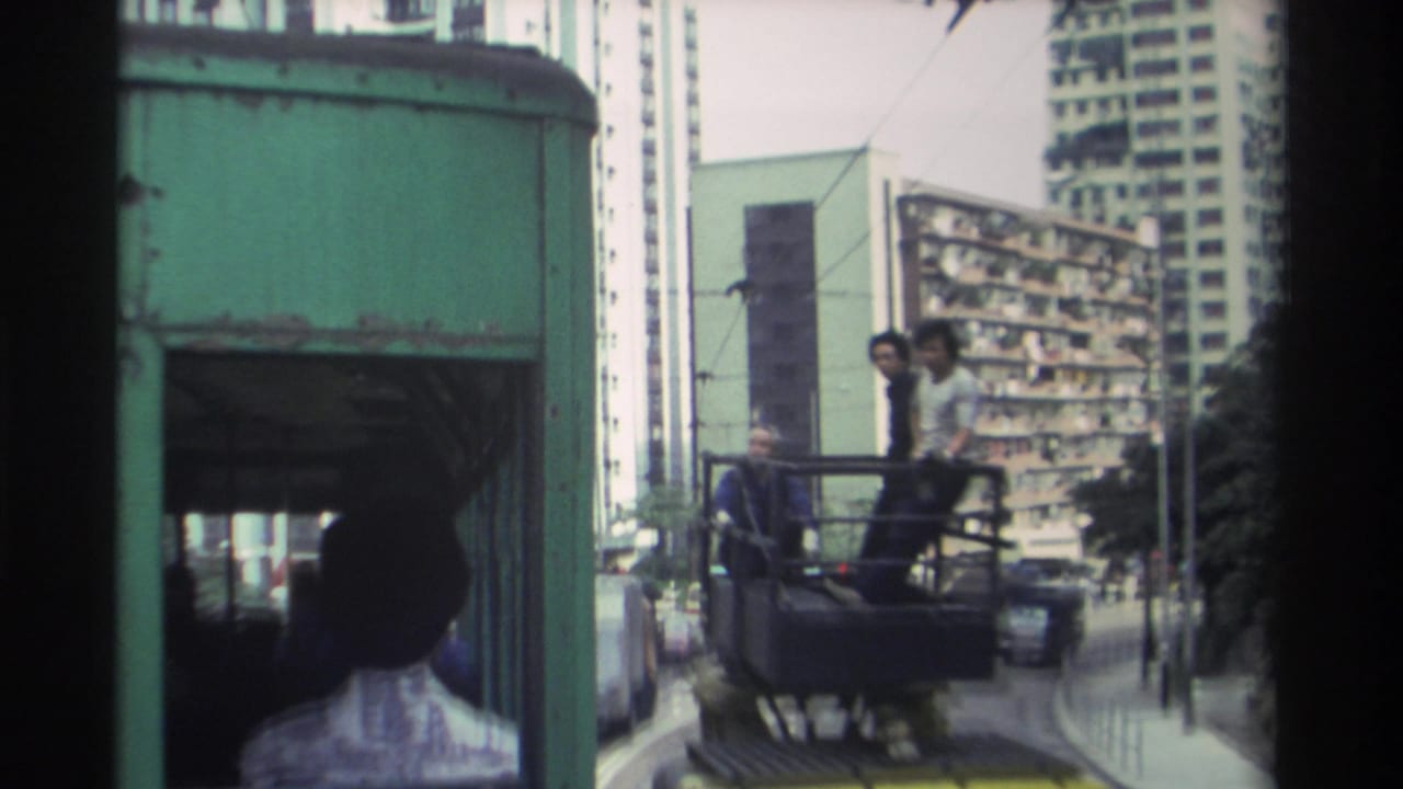 Two men on an open platform seen through the green rear structure of a tram, with dense high-rise apartment buildings visible behind.