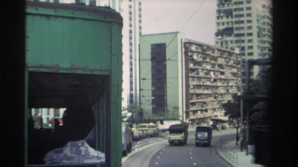 Tram's-eye view along a wide Hong Kong road circa 1982, dense slab-block residential towers right, period vehicles ahead on curving road under overcast sky.