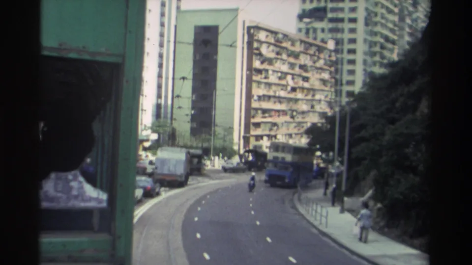 Moving tram shot of a Hong Kong street circa 1982 showing dense residential towers, a double-decker bus, a commercial van, and a motorcyclist below.