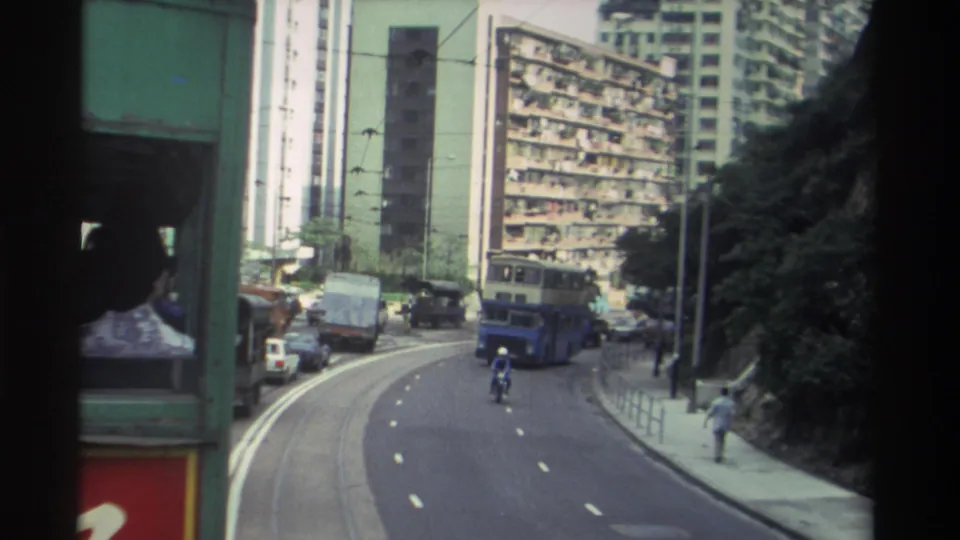 Final frame from a Hong Kong tram circa 1982: road curving right, double-decker bus, motorcyclist, wooded hillside, and partial red-yellow signage at lower frame edge.