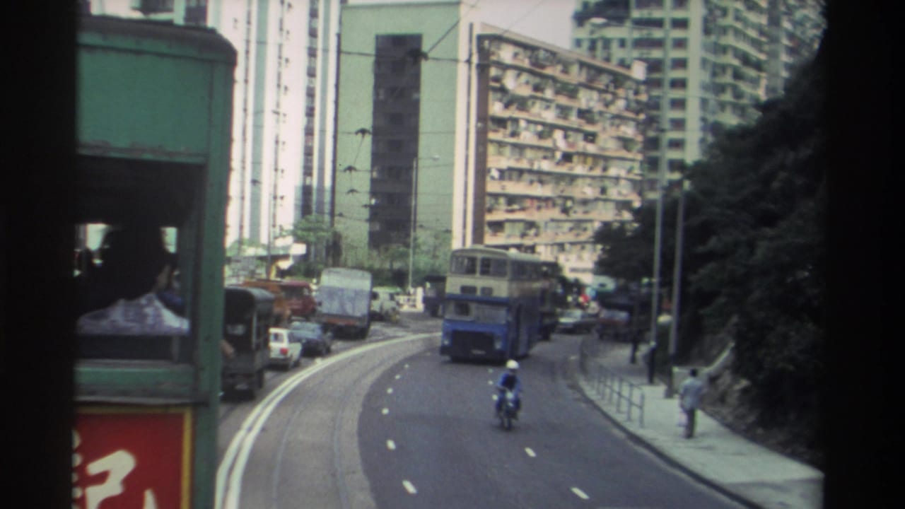 Shot from a moving tram on a curved Hong Kong road, c. 1982 — blue double-decker bus and helmeted motorcyclist in foreground, densely packed apartment towers behind.