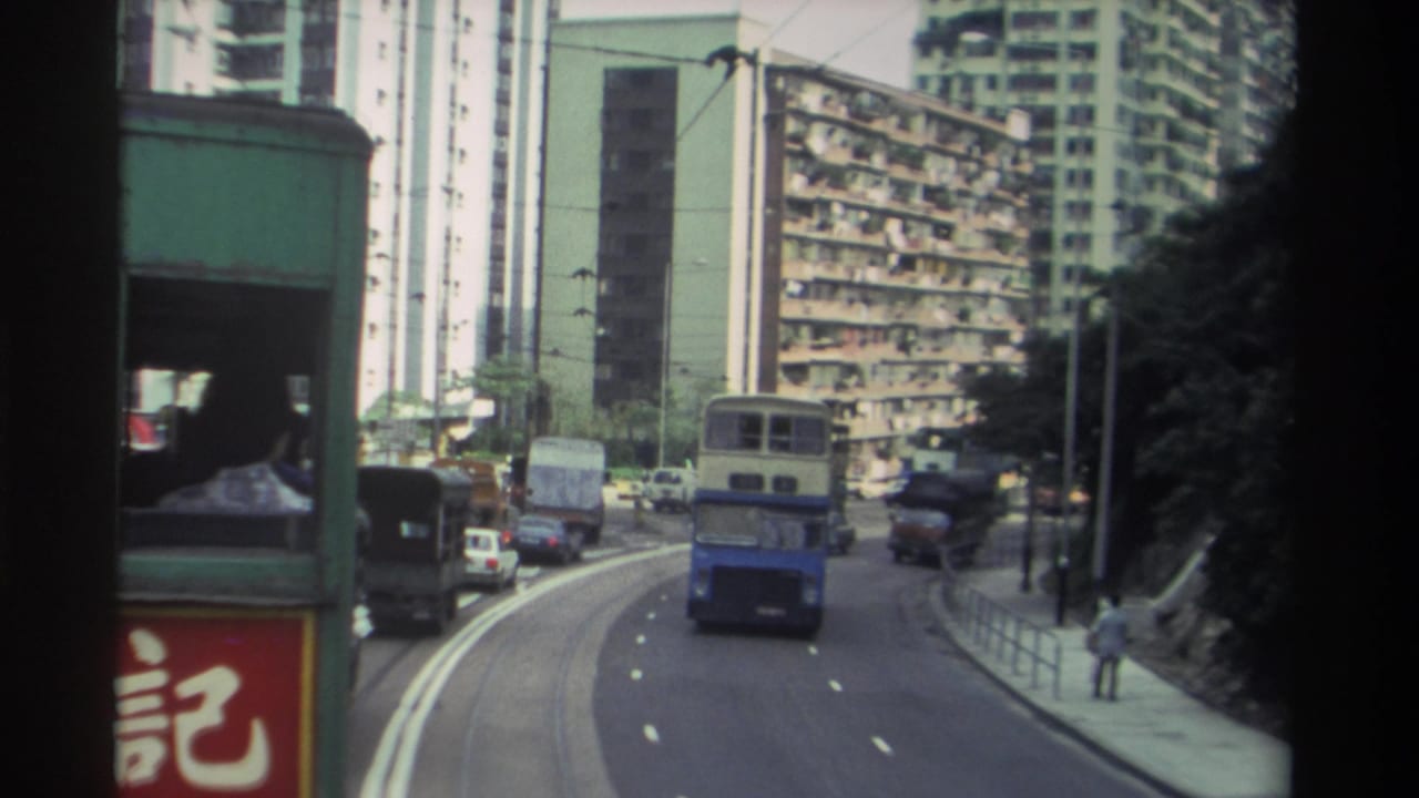 Tram-POV of curved Hong Kong road, double-decker bus ahead, pedestrian at roadside, high-rise apartment towers in background.