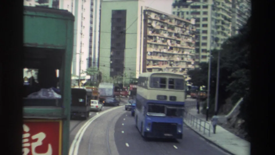 Blue-and-cream double-decker bus fills the foreground on a curved Hong Kong road, c. 1982, with dense residential towers and red Chinese-character signage flanking the street.
