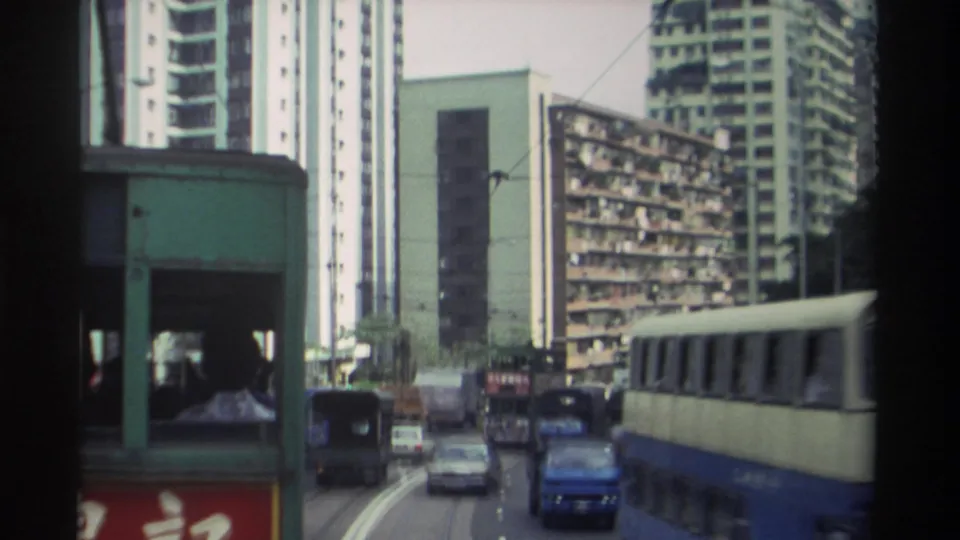 Green tram bodywork enters right of frame as camera reveals wider Hong Kong streetscape, c. 1982 — blue vehicles, dense apartment towers, and Chinese commercial signage visible.