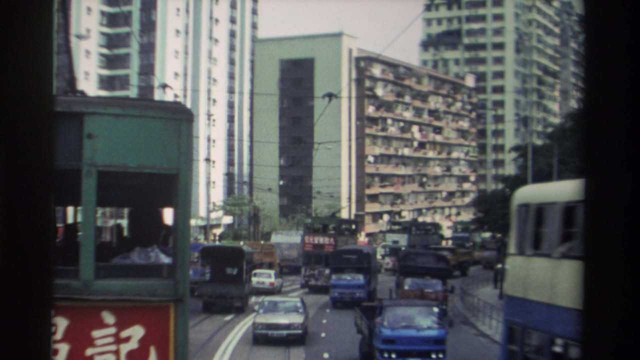 Forward view from a tram along a Hong Kong street in 1982, showing dense traffic, tall residential towers, and red Chinese shop signage.