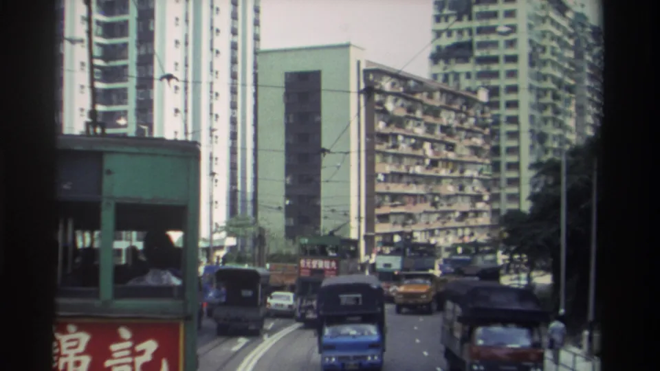 1982 Hong Kong street from tram perspective: tram visible ahead, 錦記 Chinese sign on left, dense balconied housing blocks, overhead wires, and a background pedestrian.