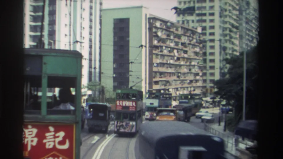 1982 Hong Kong street with a double-decker tram carrying Chinese text advertising, red 錦記 shop sign dominating the left frame, and densely balconied towers in the background.