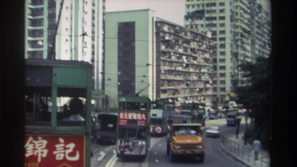 1982 Hong Kong tram corridor: yellow car prominent in mid-ground, 錦記 signage on left, double-decker tram ahead, balconied apartment blocks, and a pedestrian on the far right.