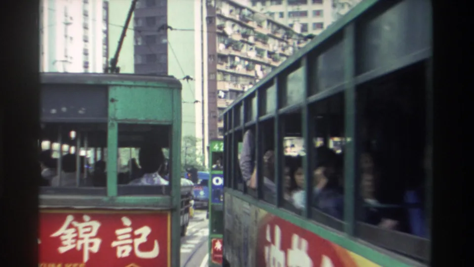 The green side panel of a Hong Kong double-decker tram sweeps past the camera; passengers are partially visible through windows, with laundry-draped high-rises behind, circa 1982.