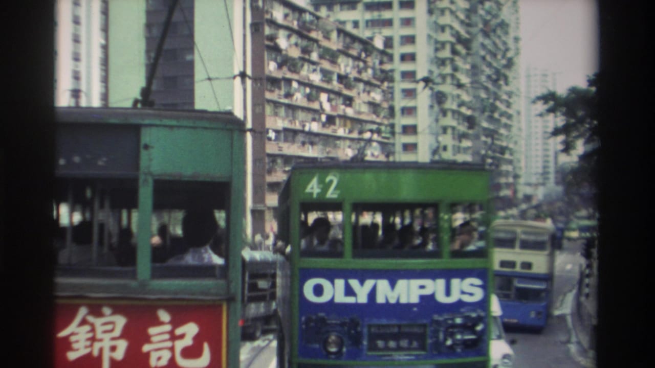 Head-on view of Hong Kong tram route 42 with a blue Olympus ad panel; red Chinese shopfront signs frame both sides; apartment towers rise behind, 1982.
