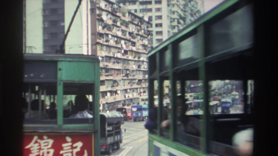 Green double-decker tram in foreground; high-density apartment blocks with laundry-draped balconies fill the background; further buses visible on the corridor, Hong Kong circa 1982.