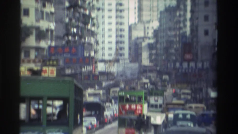 1982 Hong Kong street scene viewed through a green tram structure, showing dense traffic, multiple trams, and Chinese-character advertising on high-rise buildings.