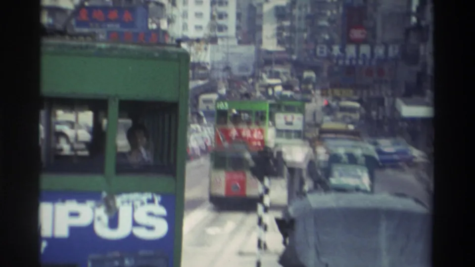 1982 Hong Kong: tram-level view with congested traffic, a partial blue billboard carrying Roman-script lettering, and Chinese-character signs on high-rise buildings.