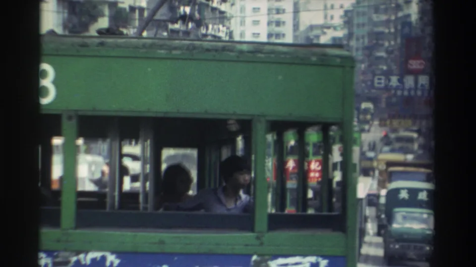 1982 Hong Kong: a passenger seen through the lower-deck window of a green tram, with Chinese advertisement signs and dense urban traffic in the background.