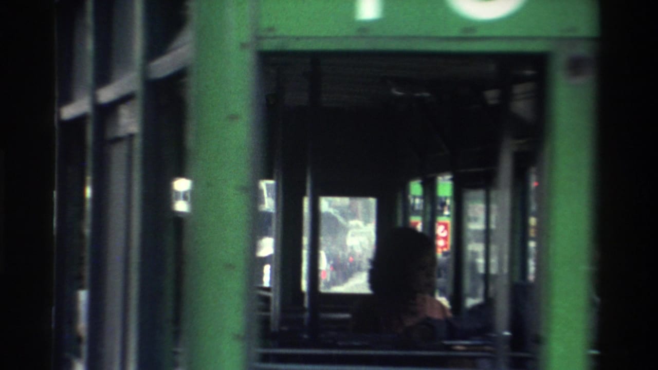 View down the aisle of a green Hong Kong double-decker bus, 1982, with a silhouetted seated passenger and a city street visible through the front windows.