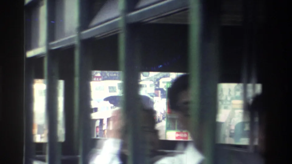 Bus passenger in a light collared shirt on a Hong Kong bus in 1982, with a busy commercial street, neon signs, and pedestrians visible through the window.