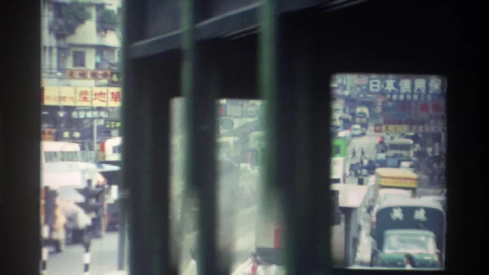 Busy Hong Kong commercial street circa 1982 framed by bus window bars, with dense Chinese-character shop signs, a large bus or truck, and a crowded pavement.