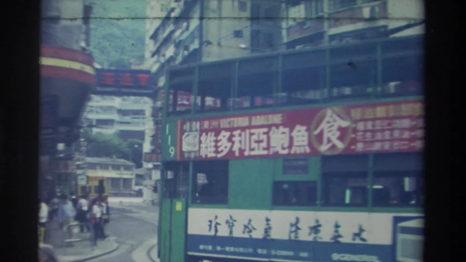 1982 Hong Kong commercial district seen from a bus window: a prominent red Victoria Abalone billboard (維多利亞鮑魚食), multi-story buildings, a white sign with Chinese text, and street-level pedestrians and vehicles.