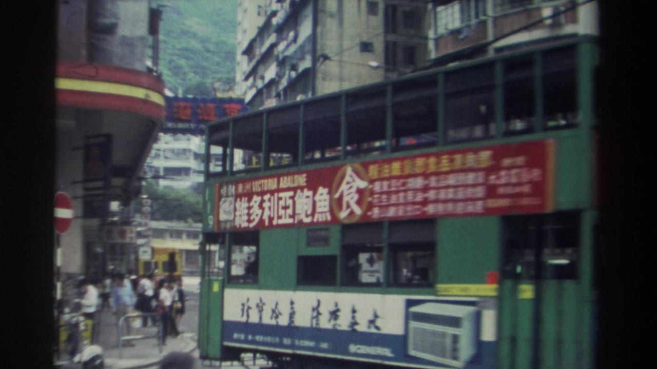 Busy Hong Kong street corner with large Chinese-character billboards, high-rise buildings, and pedestrians; green hills visible above the roofline.