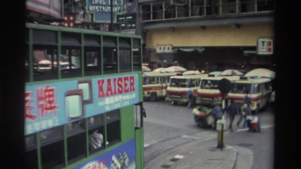 1982 Hong Kong street scene: green tram with KAISER electronics advertisement reveals a busy street behind it — red-and-cream minibuses, pedestrians, market umbrellas, and a TUNG IK RESTAURANT sign on the building facade.