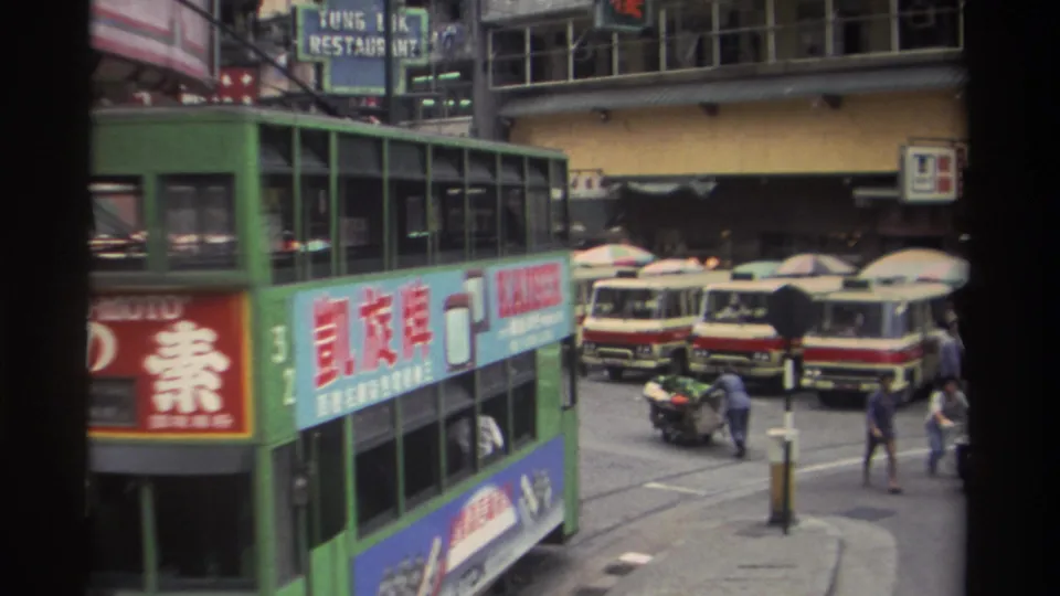 1982 Hong Kong: tram bearing KAISER advertisement alongside dense Chinese commercial signage; TUNG IK RESTAURANT sign visible, red-and-cream minibuses parked, and pedestrians moving on the pavement.