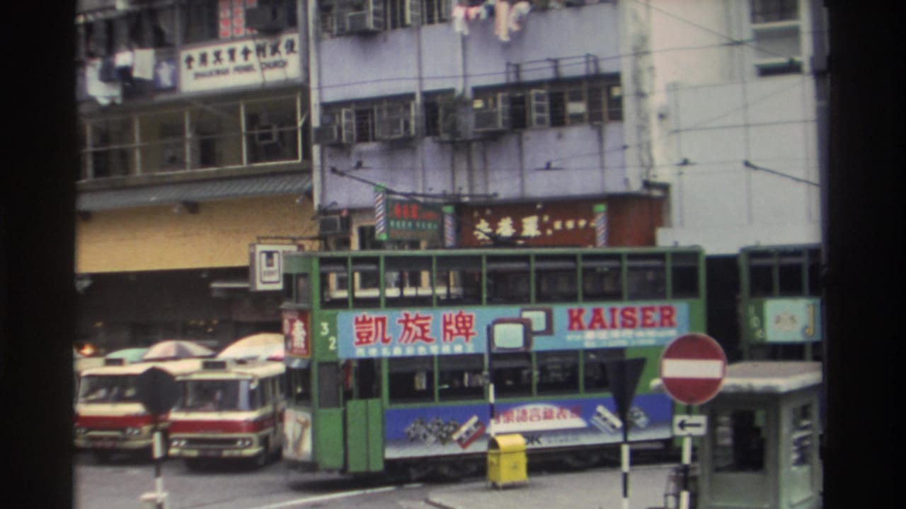 8mm footage, Hong Kong circa 1982: a green tram bearing a KAISER brand panel navigates a crowded street with Chinese commercial signage and a red double-decker bus visible left.
