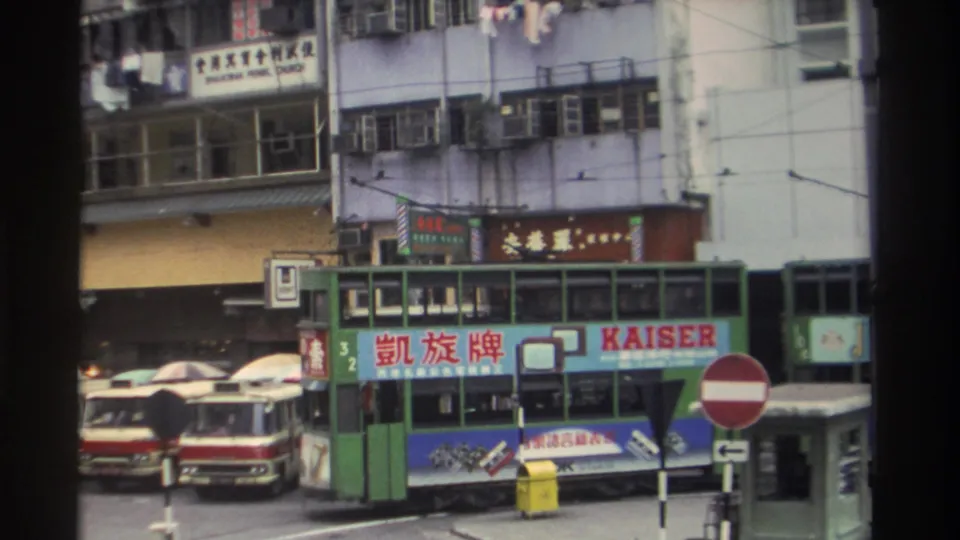 8mm footage, Hong Kong circa 1982: a green tram bearing a KAISER brand panel navigates a crowded street with Chinese commercial signage and a red double-decker bus visible left.