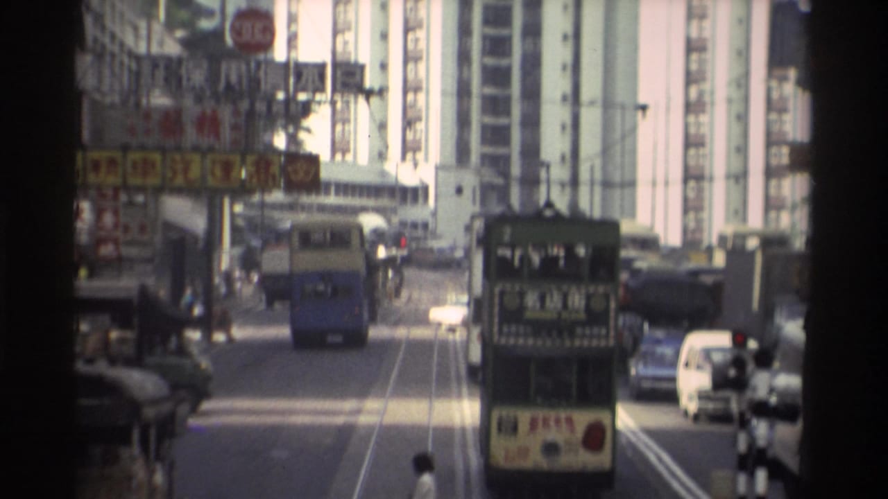 Forward-tracking 8mm view along a Hong Kong tram route circa 1982, with a blue double-decker bus and green tram ahead amid Chinese commercial building fronts and high-rises.