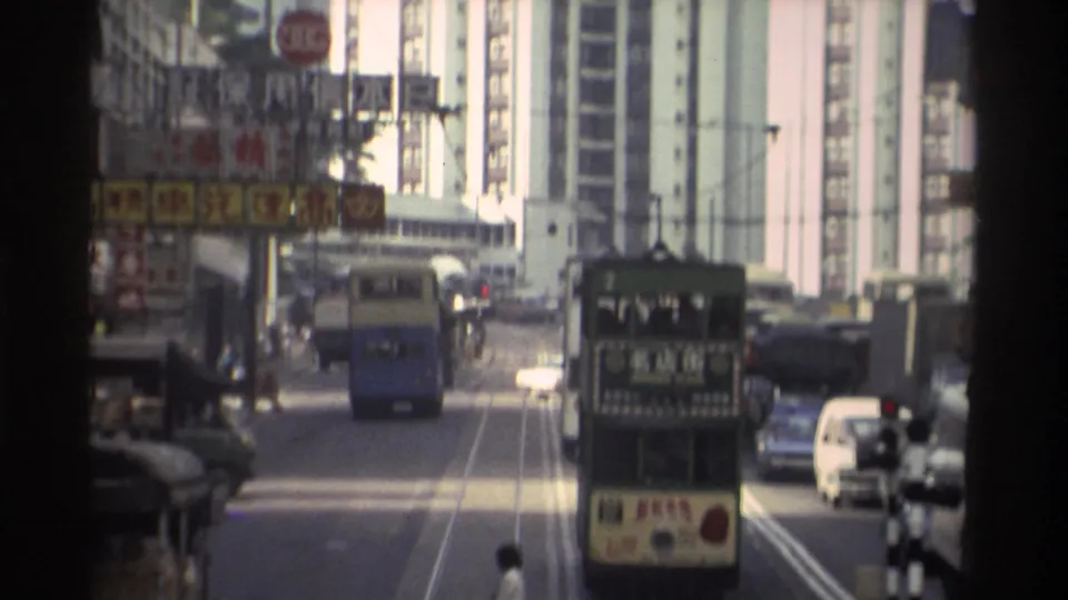 Forward-tracking 8mm view along a Hong Kong tram route circa 1982, with a blue double-decker bus and green tram ahead amid Chinese commercial building fronts and high-rises.