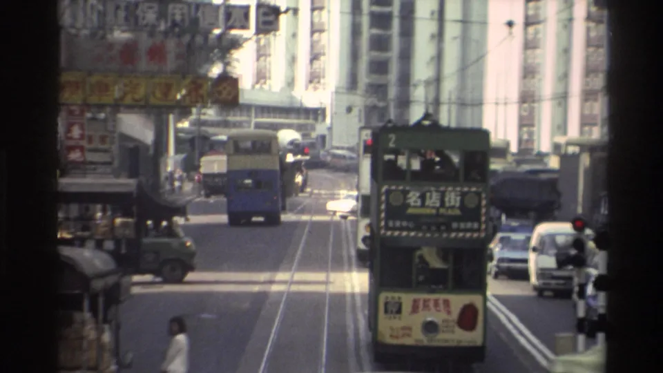 8mm Hong Kong circa 1982: a green double-decker tram on route 2 with Chinese destination characters leads mixed traffic beneath hanging commercial banner signs.