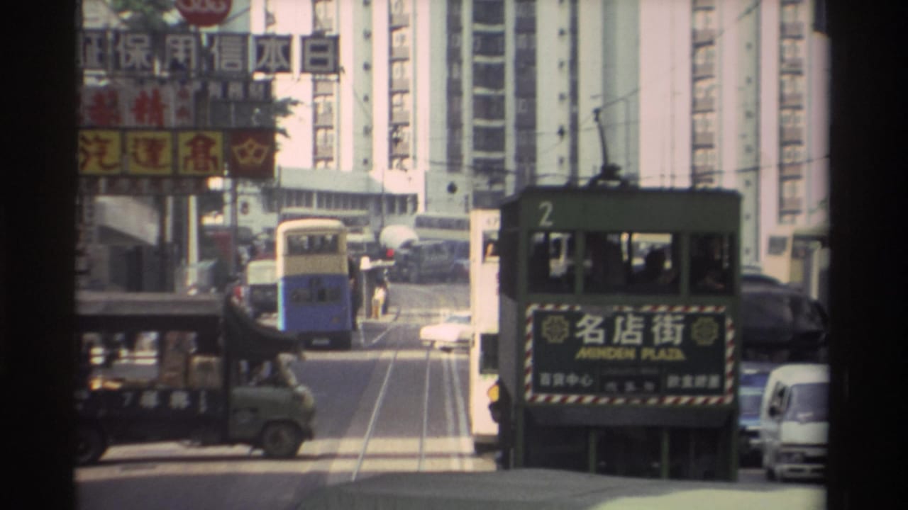 Green double-decker tram (route 2, Minden Plaza) has advanced slightly along the tram tracks; a blue bus and Chinese-language signage are visible behind it.