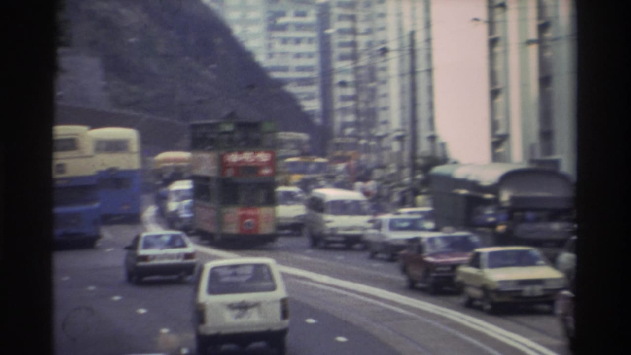Multi-lane Hong Kong road with heavy traffic including double-decker trams and buses, rocky hillside and high-rise towers in background.