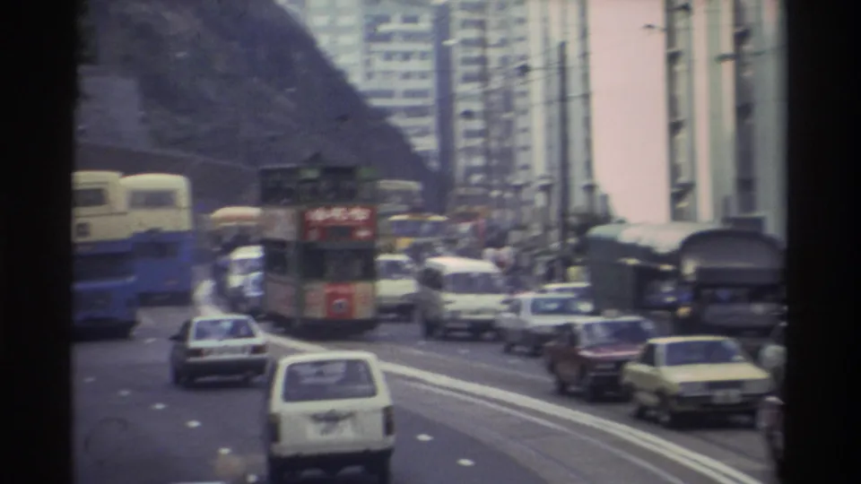 Wide shot of a busy multi-lane road in Hong Kong, circa 1982, with double-decker trams and buses among cars and vans, rocky hillside rising behind high-rise residential towers.