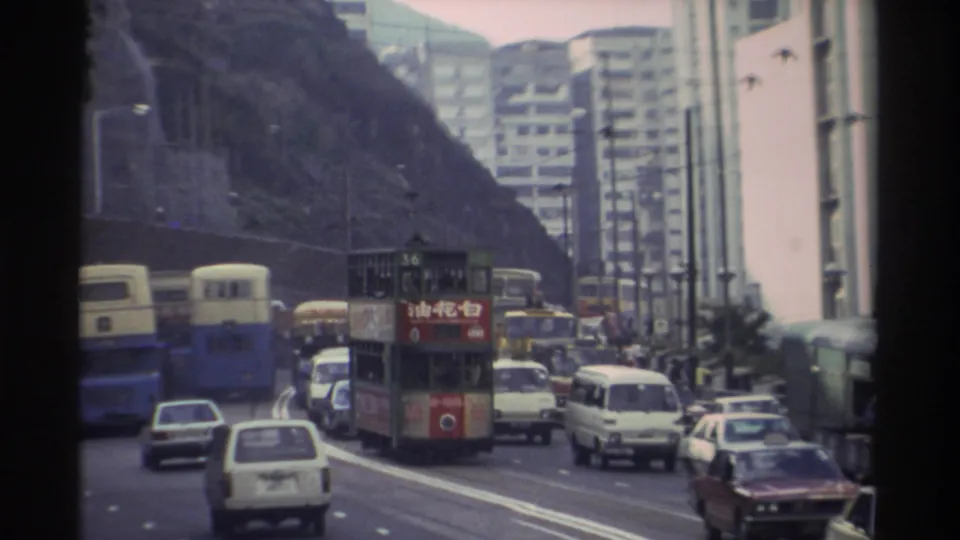 Street-level view of dense traffic on a Hong Kong urban road, circa 1982, showing double-decker trams with Chinese advertising, blue buses, and early-1980s cars with a rocky escarpment and high-rises behind.