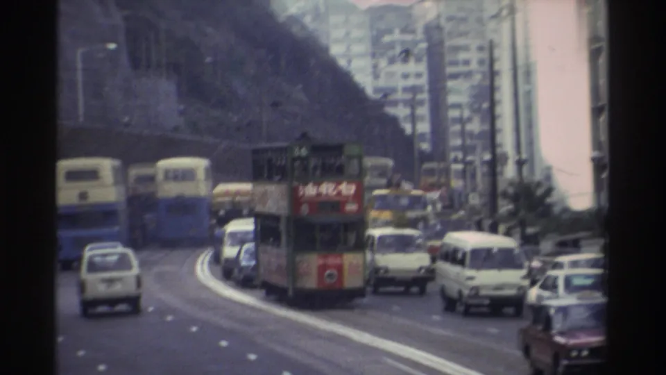 Moving-vehicle shot of a curved Hong Kong road, circa 1982, with double-decker trams displaying Chinese text and red advertising panels, buses, and period cars, framed by rocky hillside and tower blocks.