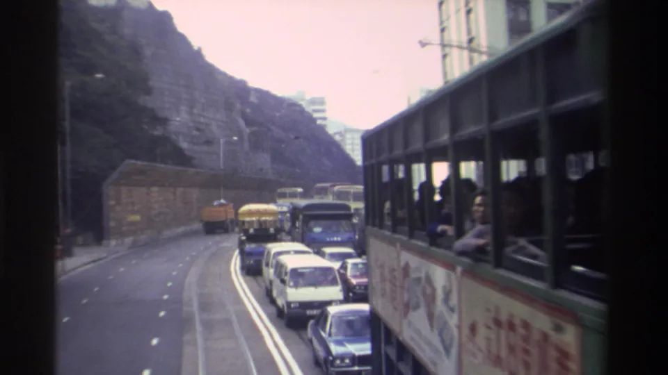 Interior vantage from a Hong Kong tram looking sideways, steep rocky hillside to the right, adjacent vehicle with advertisement visible, circa 1982, 8mm film.