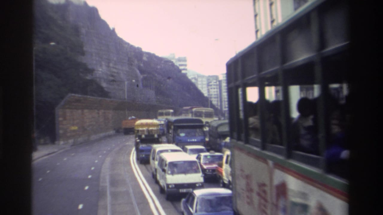 Multi-lane Hong Kong road with heavy traffic including vans, trucks, and a large bus, shot from an elevated position with a rocky hillside in the background.