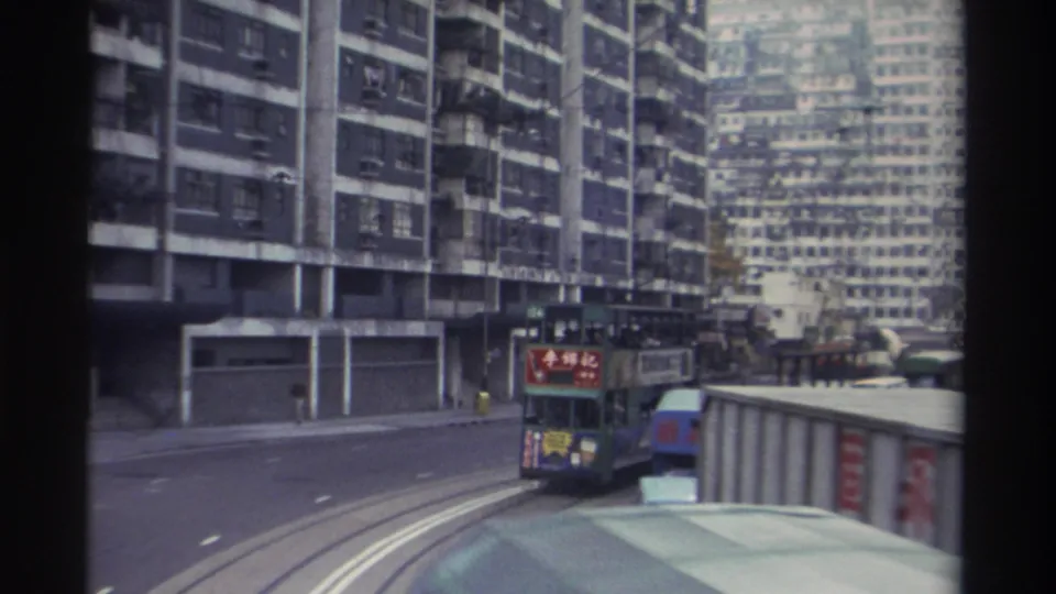 Green double-decker Hong Kong tram bearing Chinese brand advertising travels along a wide street lined with tall concrete residential estates, circa 1982.
