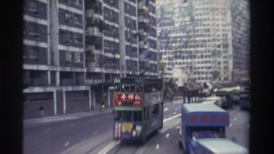 Green double-decker tram bearing Chinese advertising dominates this 1982 Hong Kong street scene, with concrete housing estates and a blue bus visible alongside, shot from an elevated vantage.