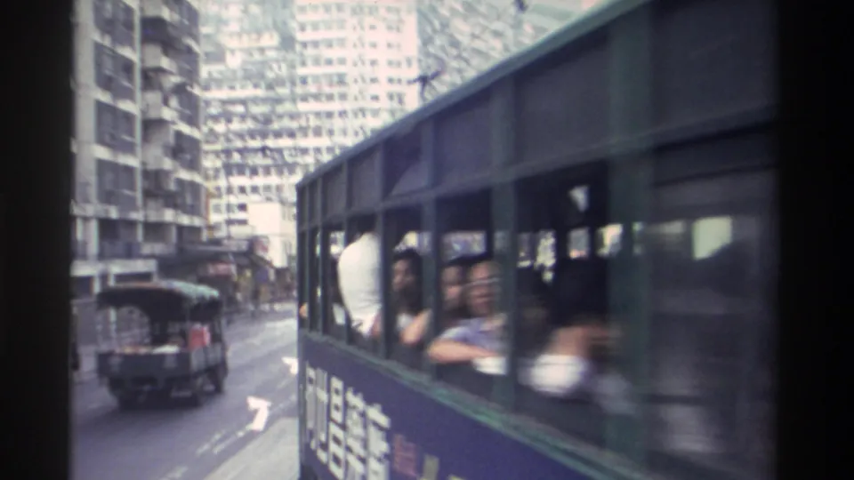 Handheld 8mm shot from alongside a moving Hong Kong tram, passengers visible through upper-deck windows, dense residential towers filling the background, circa 1982.