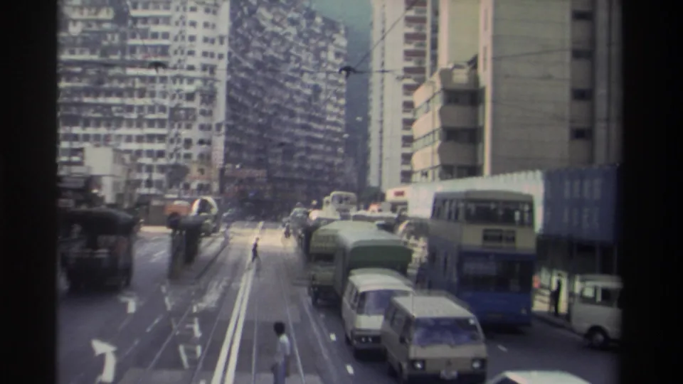 Wide 8mm view down a Hong Kong tram street showing multiple double-decker vehicles, a pedestrian on the road, overhead wires, and a dense wall of high-rise towers, circa 1982.